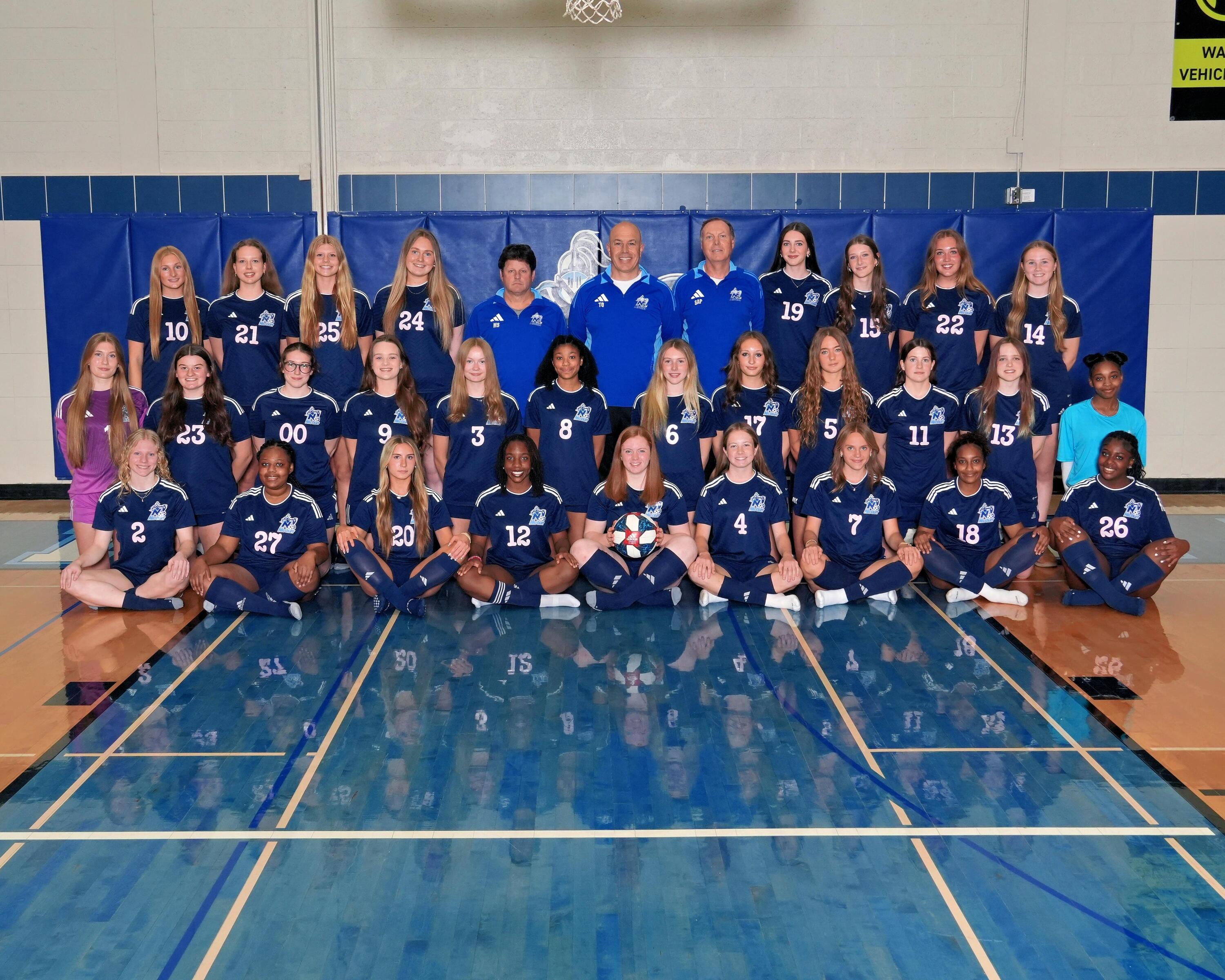 A women's soccer team in blue uniforms poses on a court with a blue backdrop. The coach stands in the center.