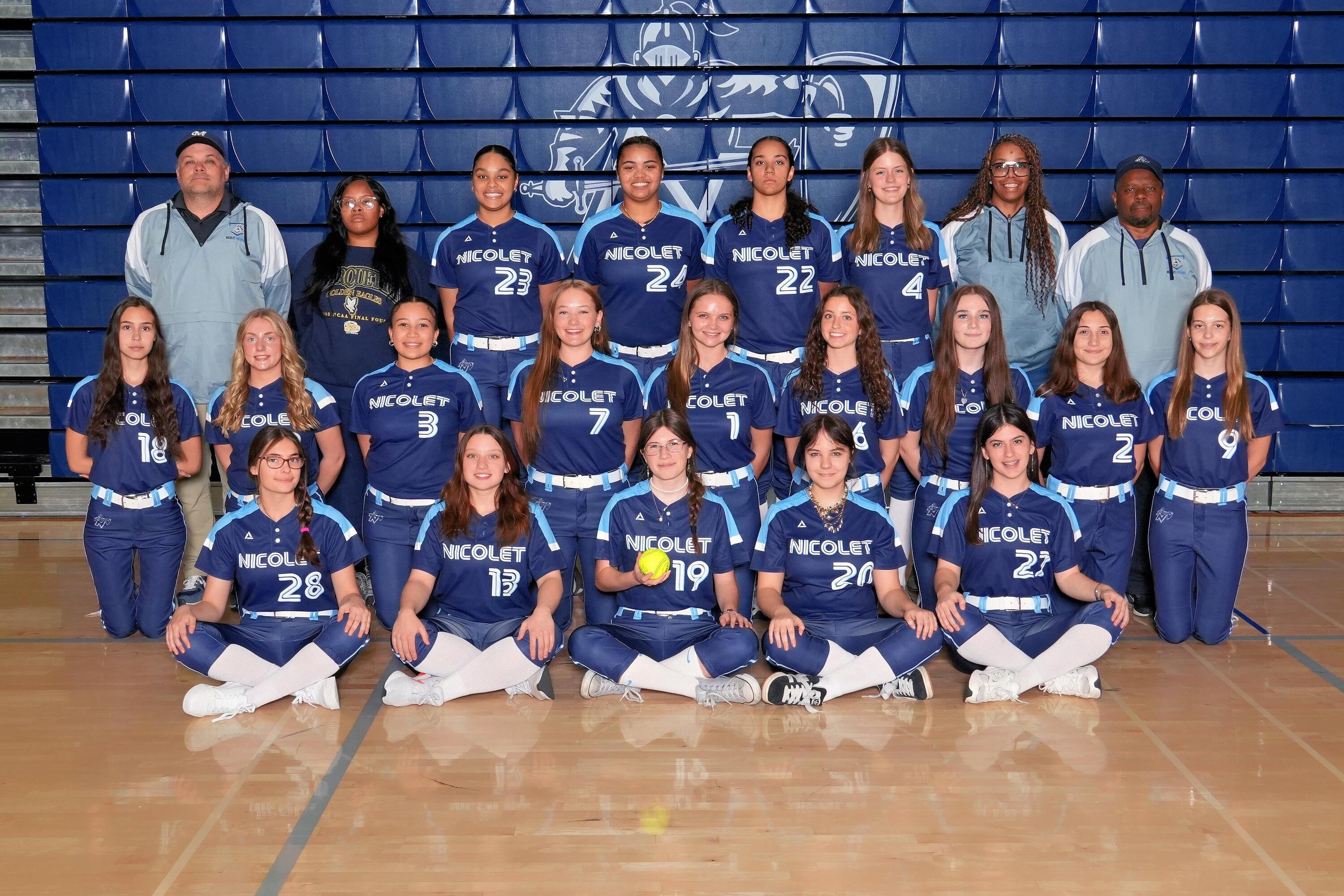 A group of athletes in blue uniforms and numbers pose on a basketball court with blue bleachers in the background.
