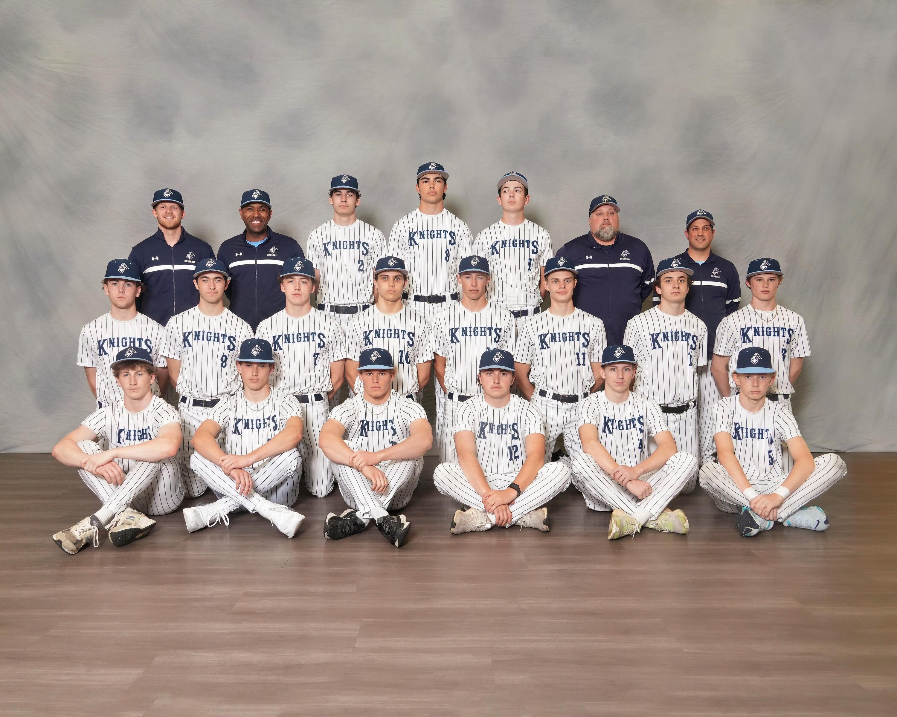 A sports team in white uniforms and caps poses against a gray backdrop with wooden flooring.