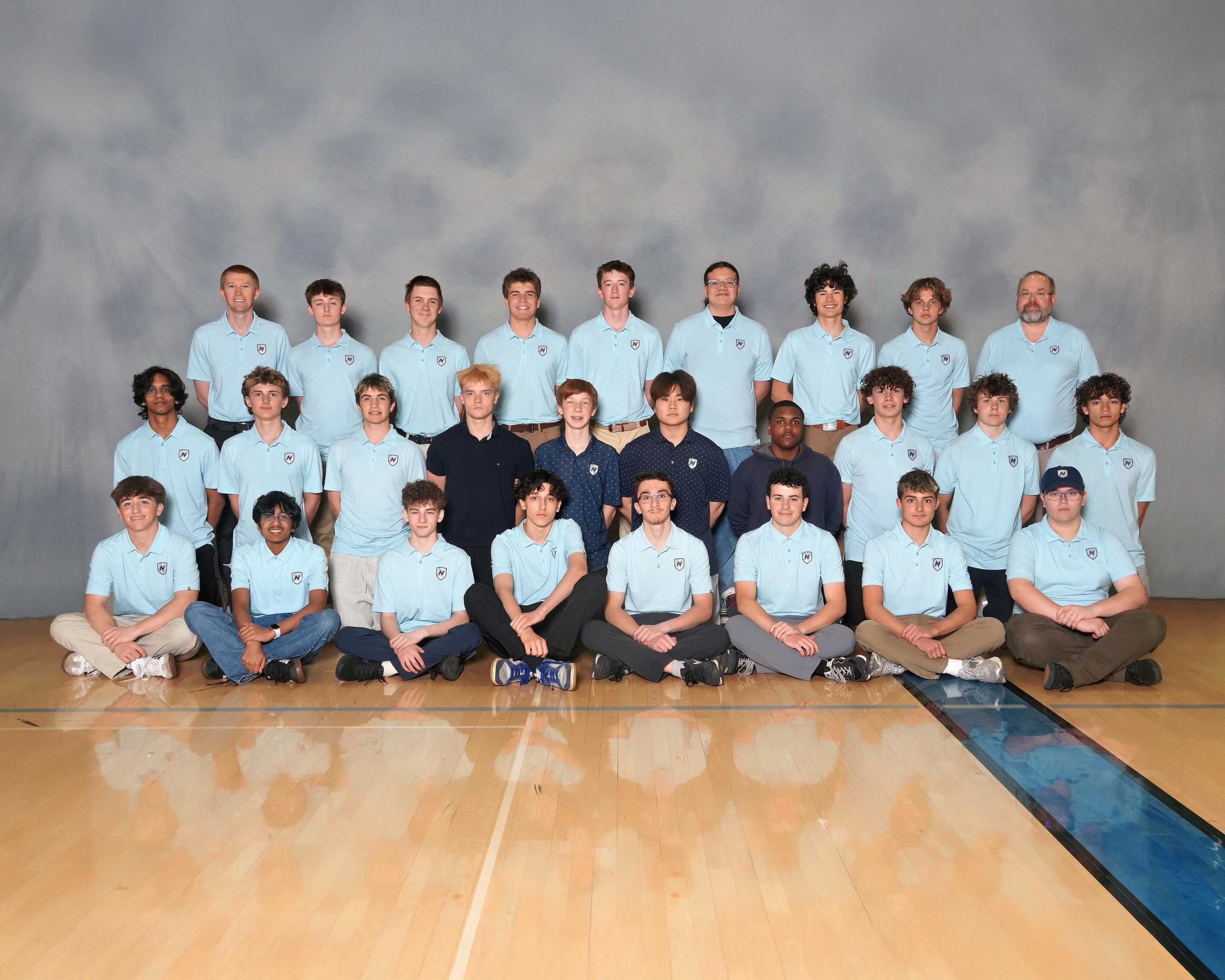 A group of individuals wearing blue shirts and sneakers, posing in a gymnasium with a wooden floor and a blue line.