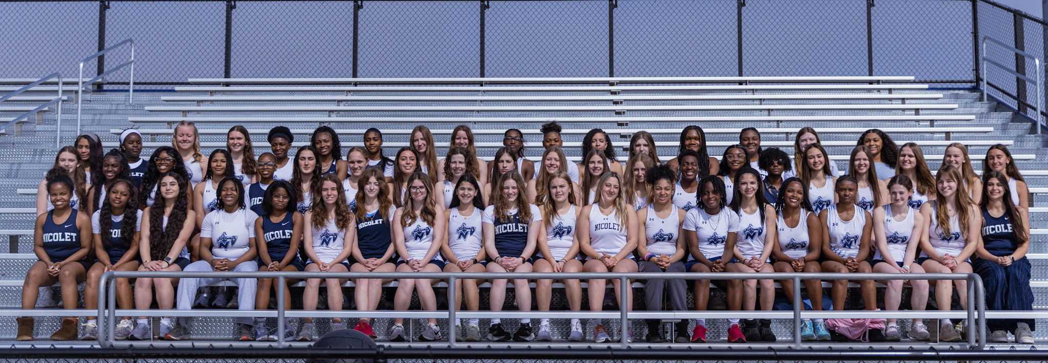 Group photo of sports team with white and blue uniforms sitting and standing in rows on stadium bleachers.