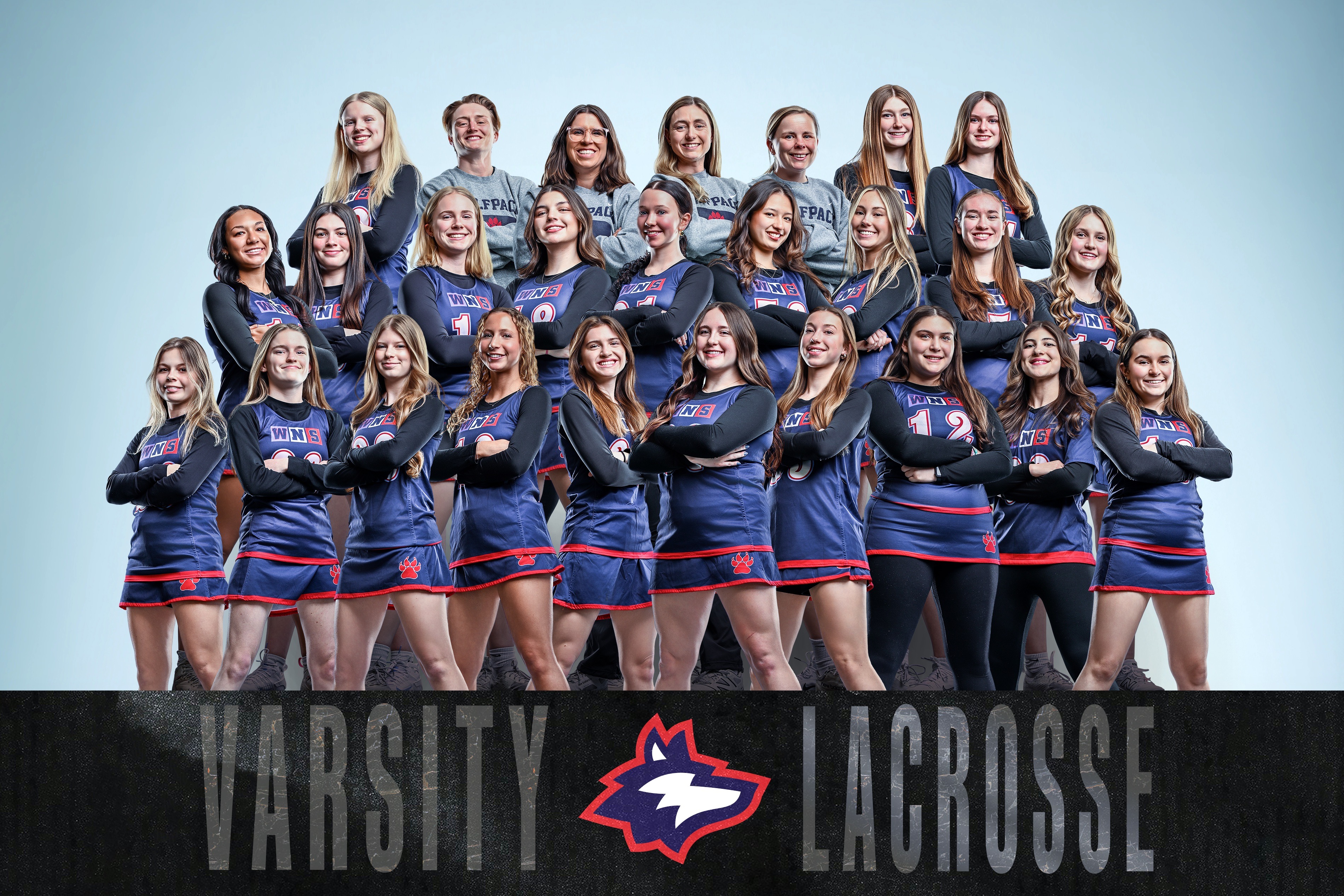 A group of female athletes in blue uniforms pose for a team photo with the words "Varsity Lacrosse" below.