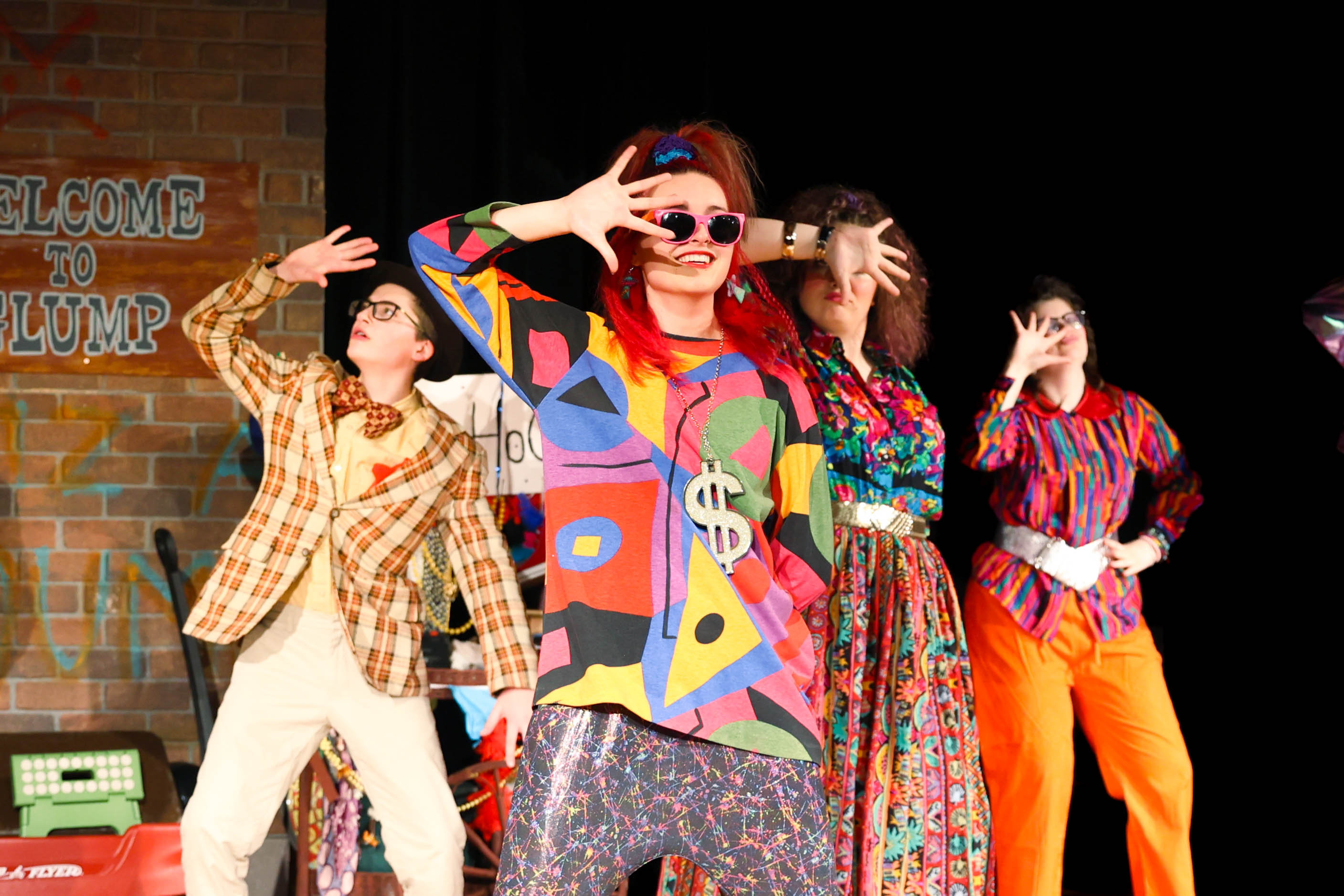 Four performers on stage. One wears a colorful, abstract outfit. Three others wear patterned, 1970s-style clothing.