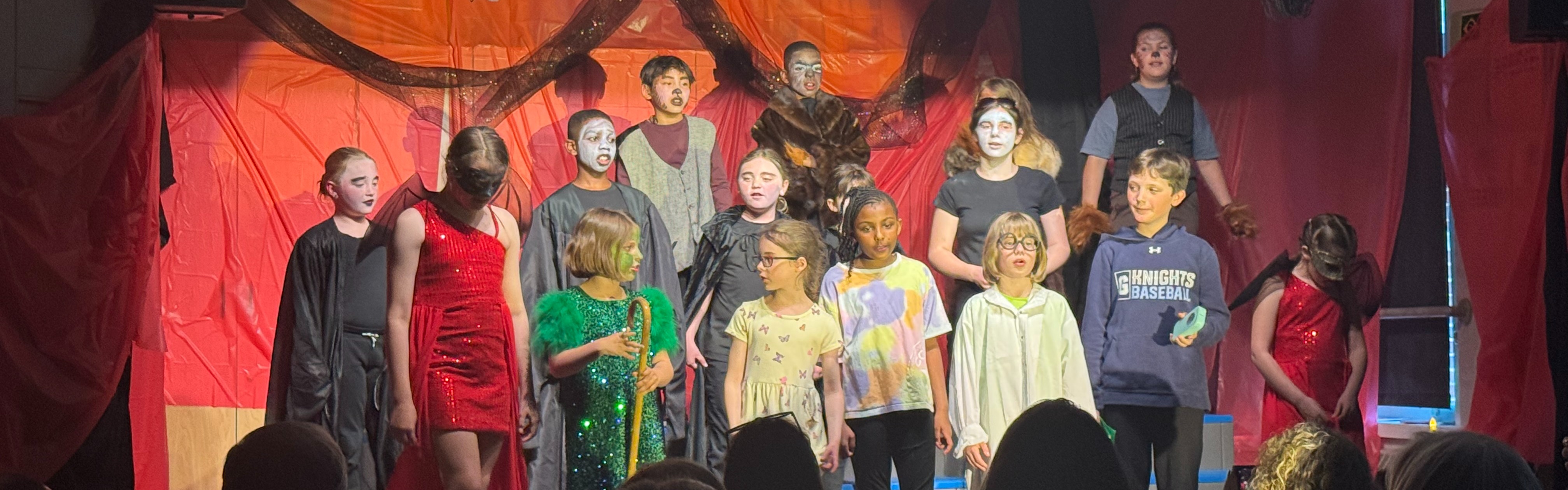 A group of children in costumes stand on a stage, with an audience watching. The stage has a red backdrop and lighting.