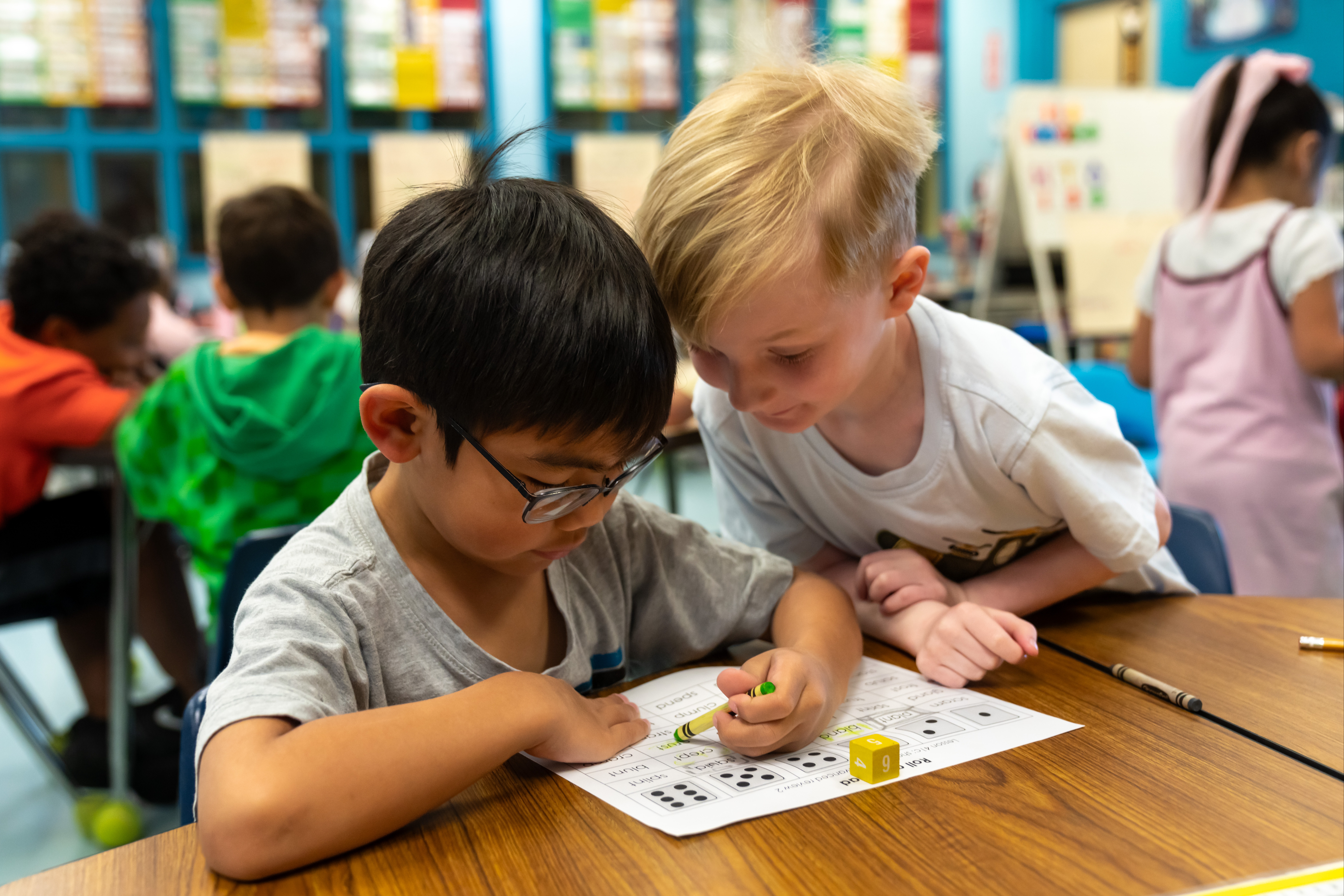 Two boys sit at a table, one with glasses, and both appear to be working on a worksheet.