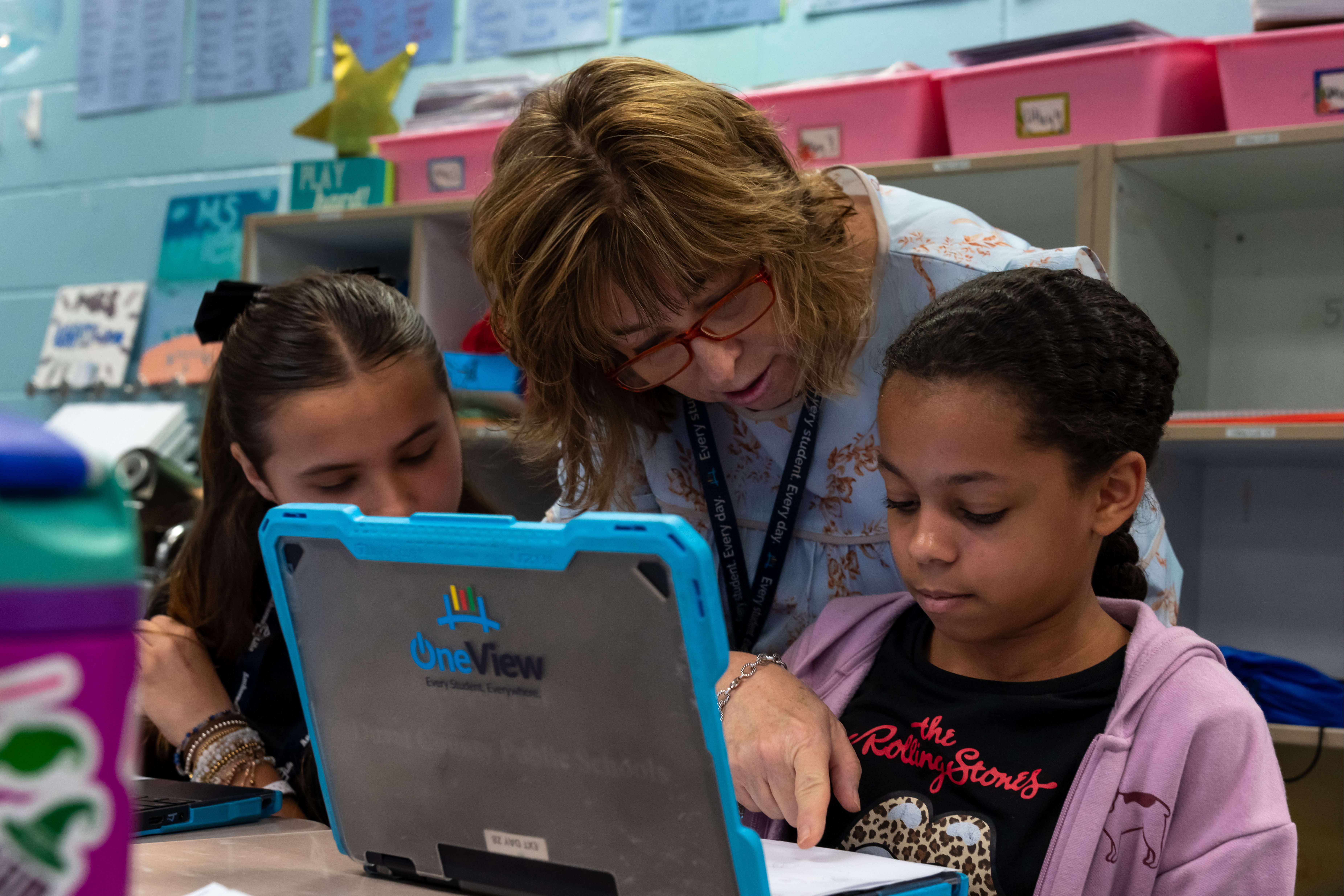 A woman helps two young girls with a laptop in a classroom. One girl wears a black shirt, the other a pink jacket.