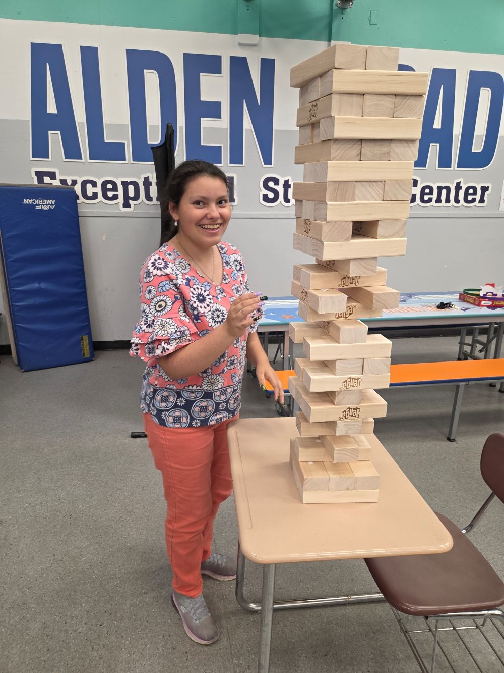 A woman stands in a gym, smiling beside a tall stack of wooden blocks on a table.