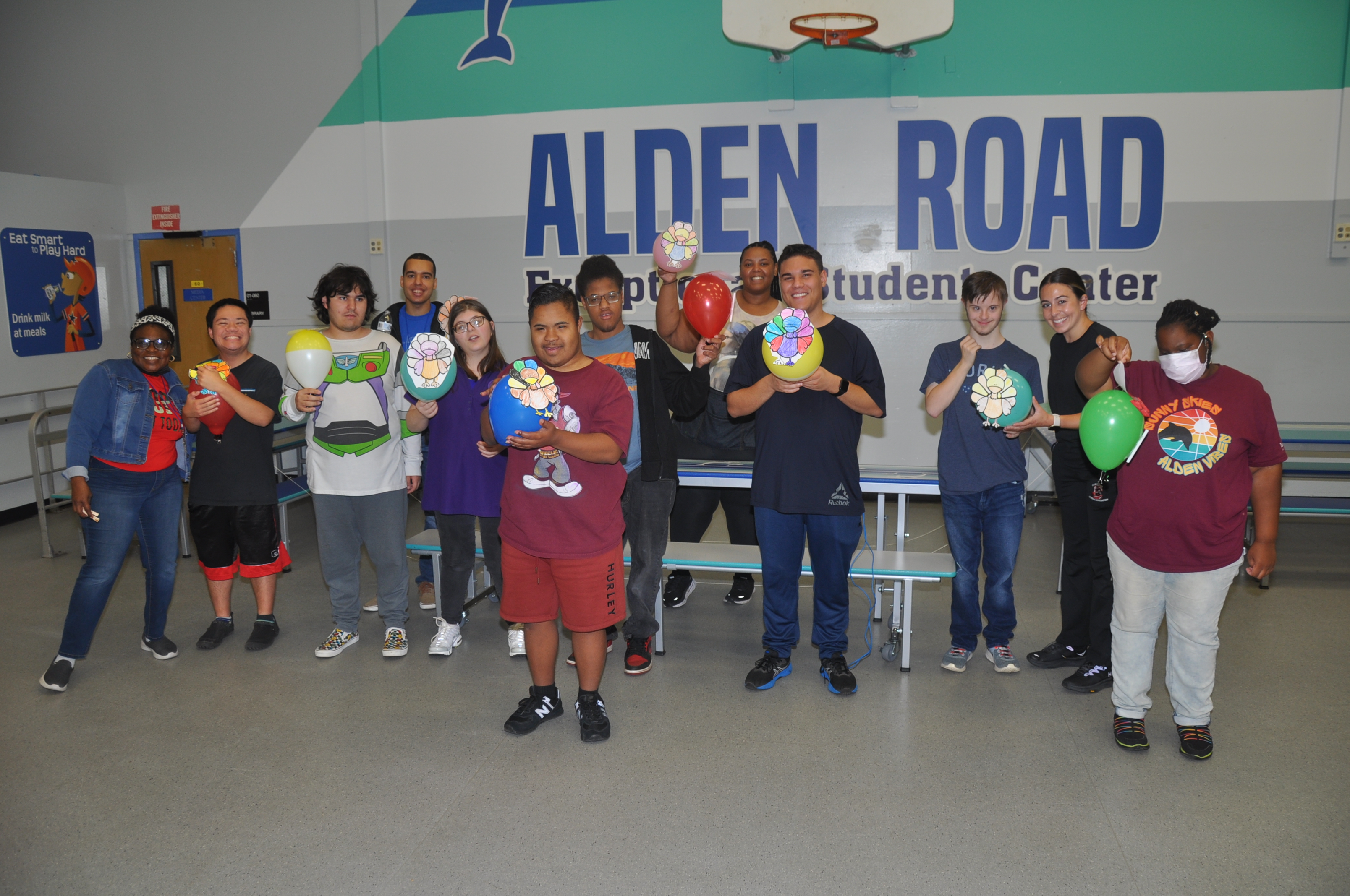 People stand in a group, smiling and holding balloons, in a room with a wall that reads Alden Road.