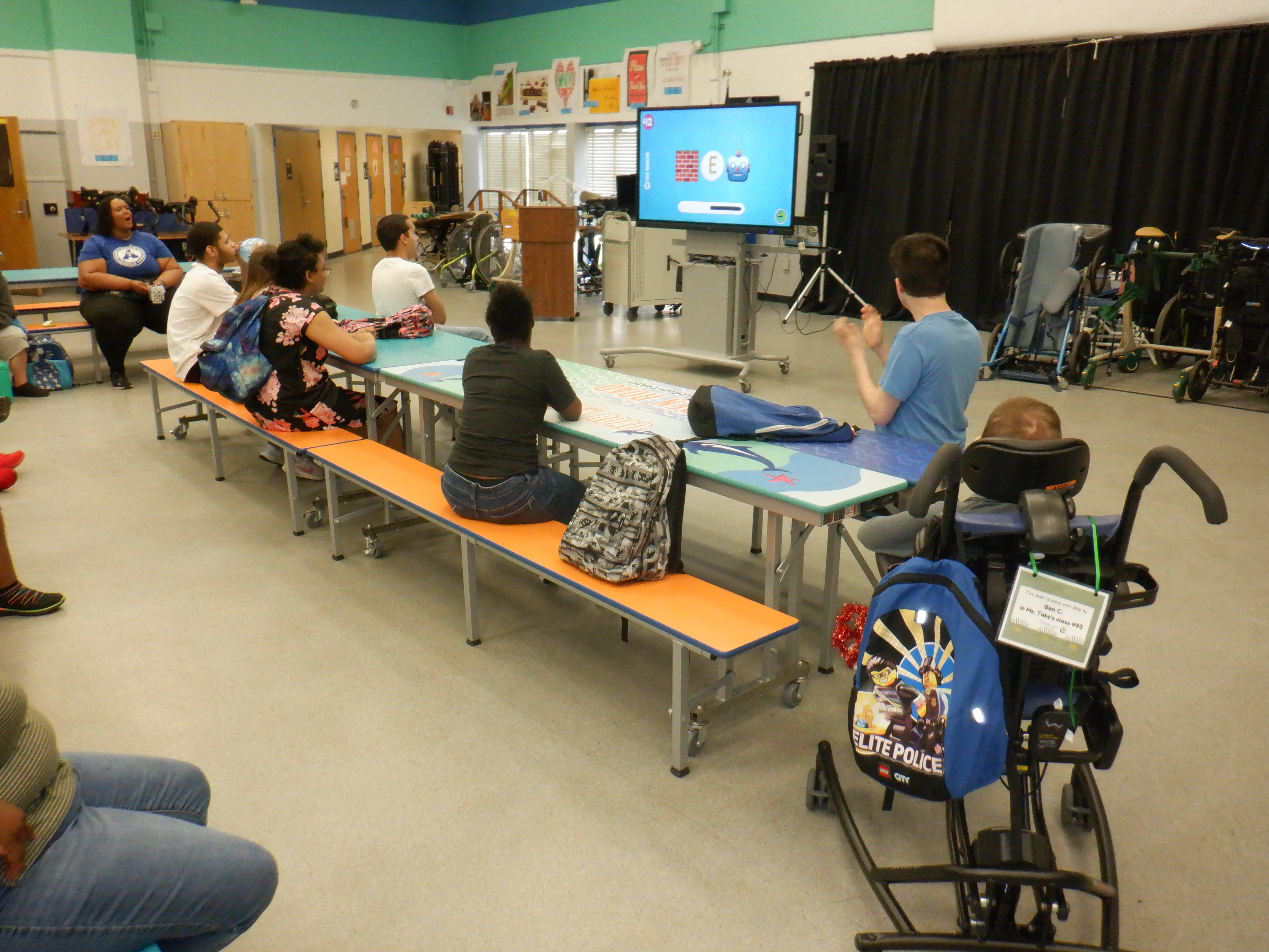 Group in a room with a large screen in the background. People sit on benches and a person sits in a wheelchair.