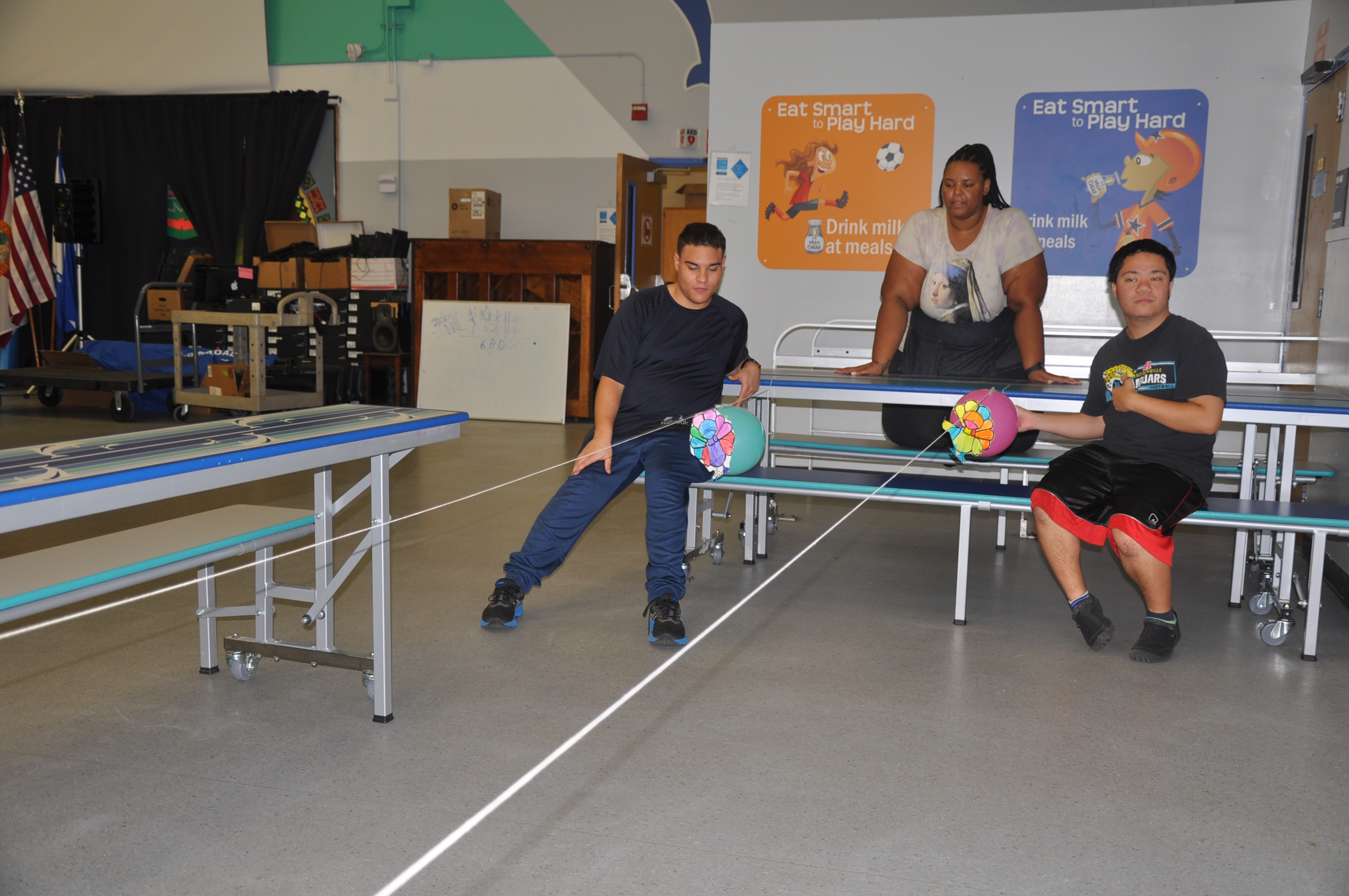 Three students seated at a table holding strings with toys attached. A wall with posters is in the background.
