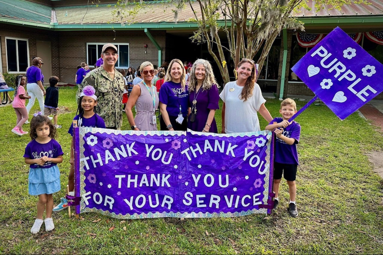 Group of adults and students standing behind a purple banner that says, "Thank you.  Thank you. Thank you for your service."