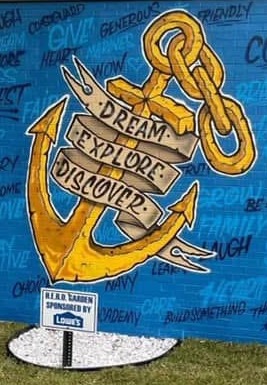 A mural on a building wall with an anchor, "Dream Explore Discover" text. Words like "cool", "funny", "champion" are painted in blue. A Lowe's sign is in front of the mural.