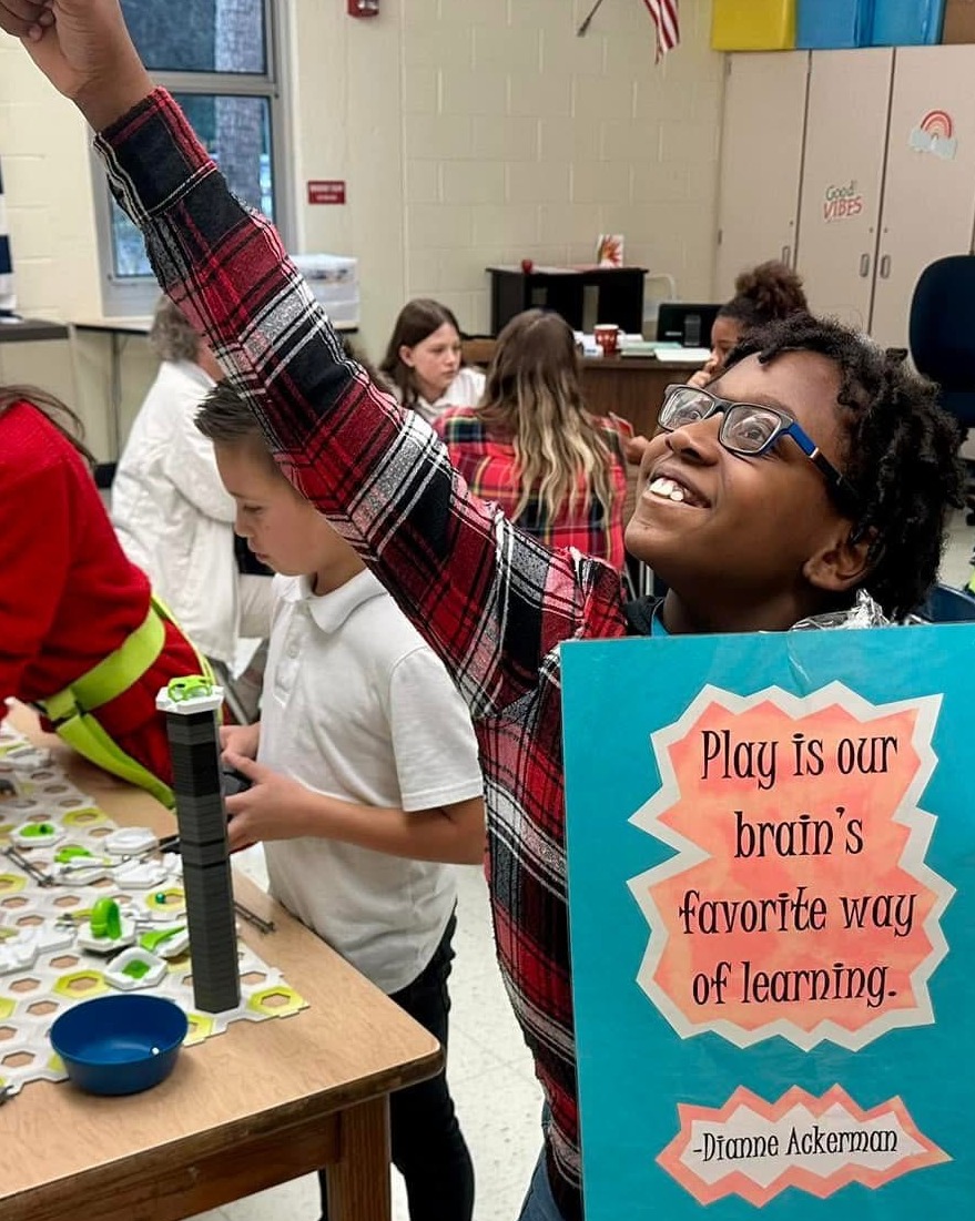 A classroom with kids. A child holds a blue sign that reads "Play is our brain's favorite way of learning".