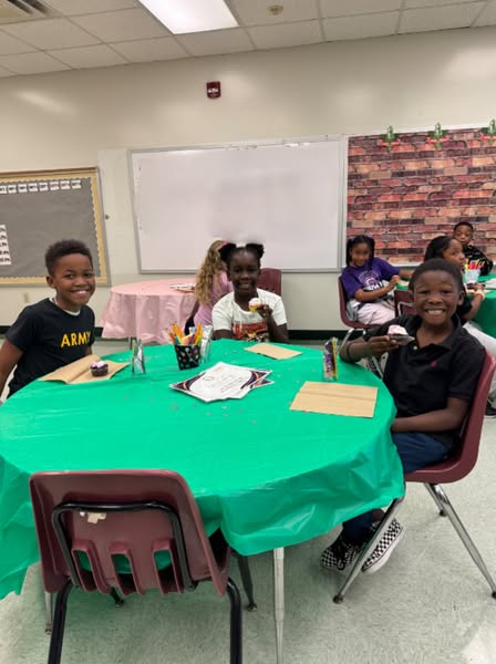Four children sitting at a table with green covers, smiling. One wears an Army shirt. Behind them, a whiteboard and brick wall.