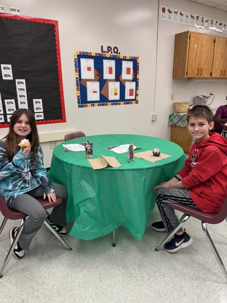 Two children at a round table, one eating a cookie. A bulletin board with squares is behind them.