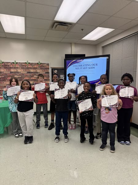 Children in a room holding certificates, standing in front of a classroom screen displaying "Celebrating Our Military Kids".