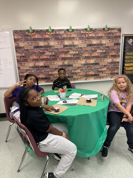 Four children seated around a green table in a room with brick-patterned wall and white board.