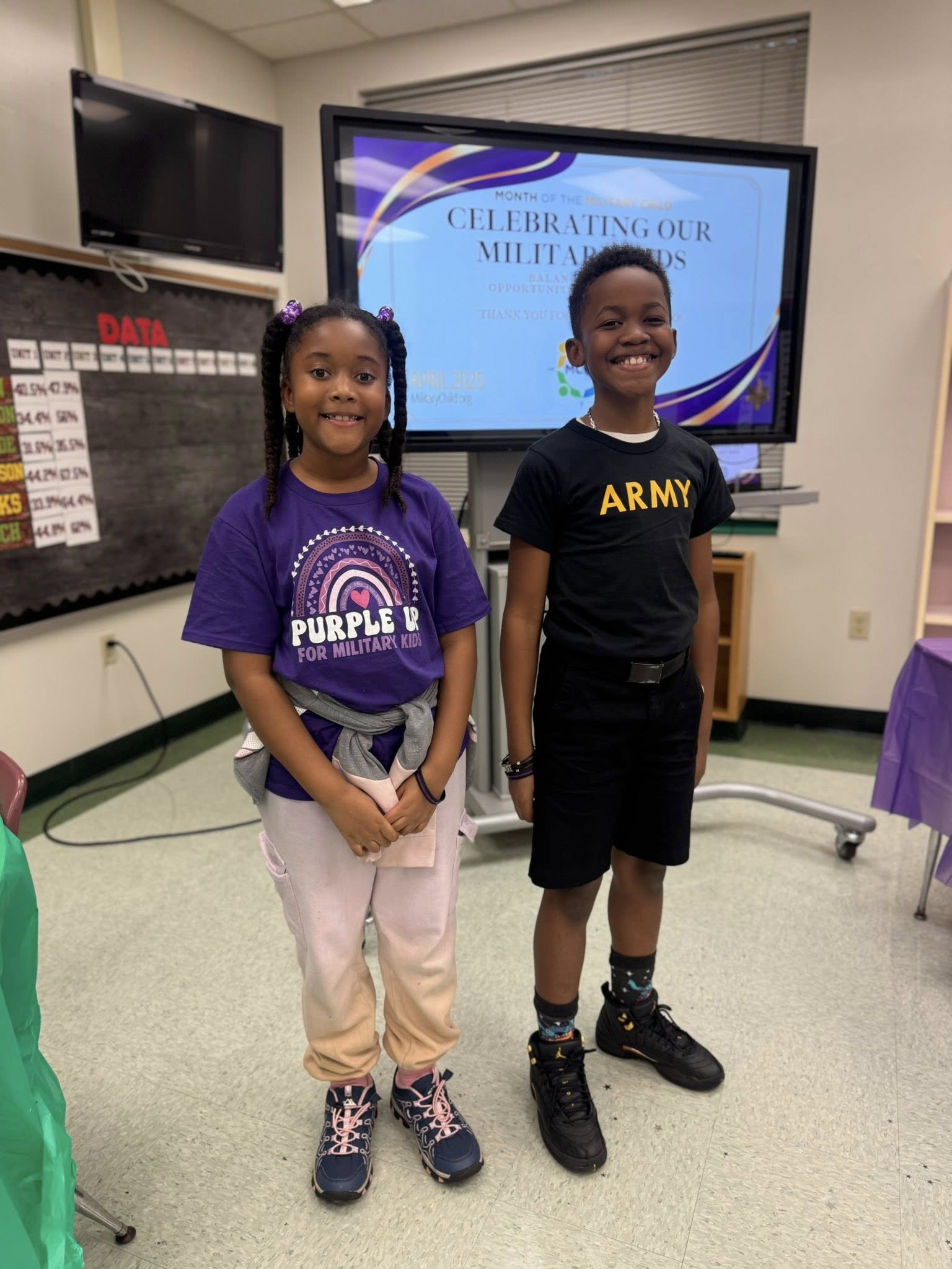 Two children in a classroom with a screen displaying "CELEBRATING OUR MILITARY HEROES". One wears a purple shirt, the other a black one.