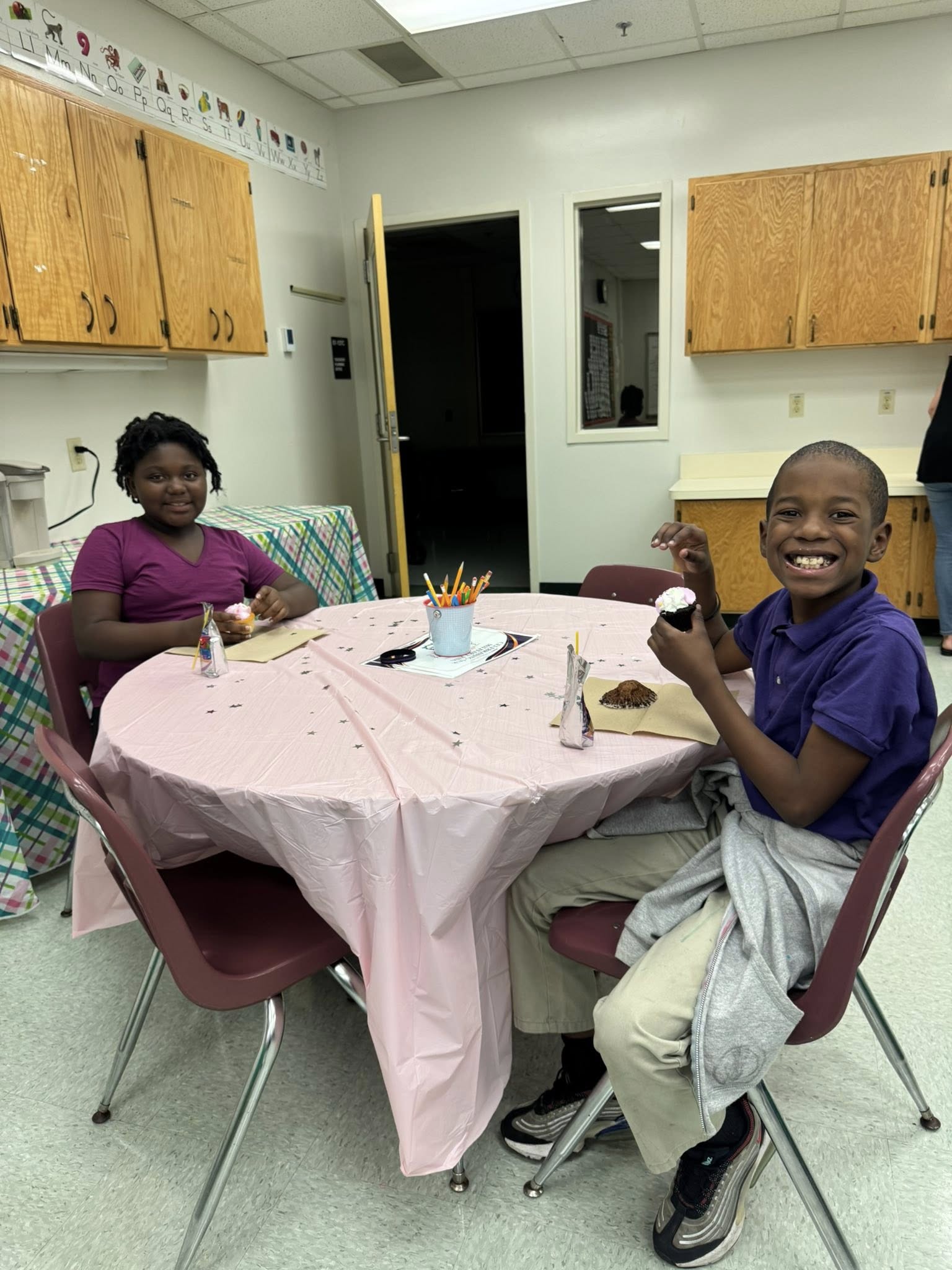 Two children seated at a table with a pink tablecloth. They hold food and utensils. Behind them, a door and wooden cabinets are visible.