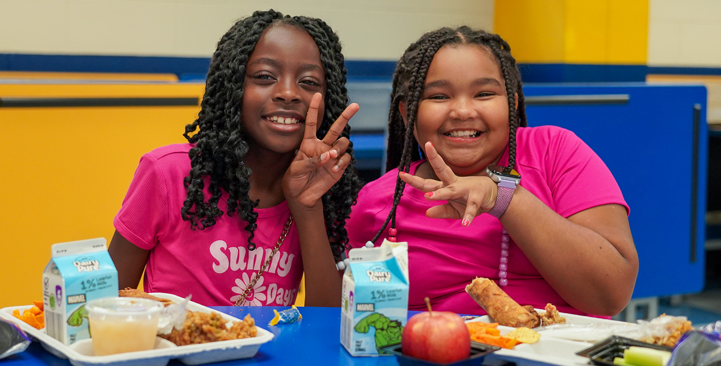 two girls eating at the lunch table giving peace signs to the camera