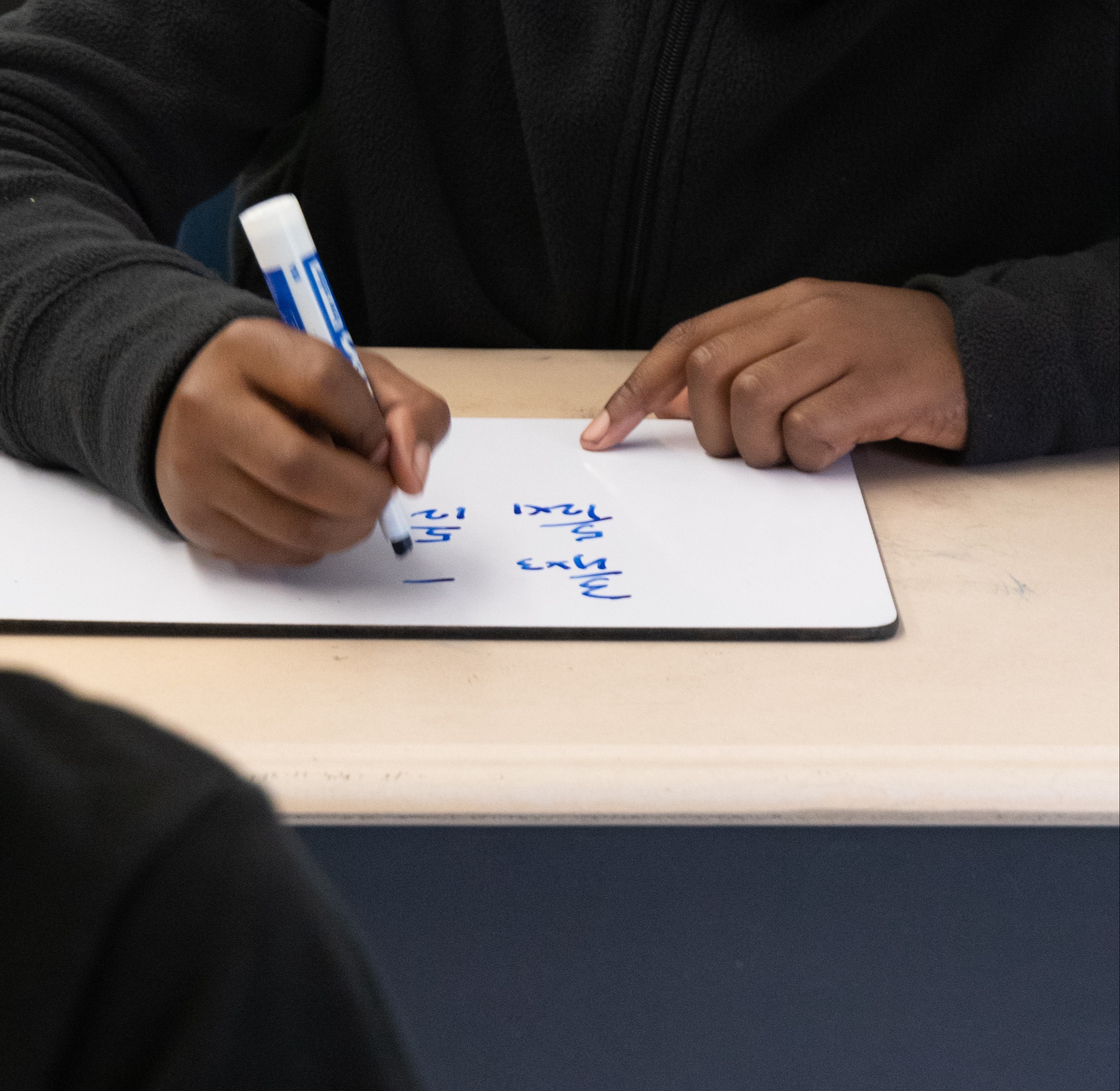 A person writes on a white board with a blue marker, seated at a light brown desk.