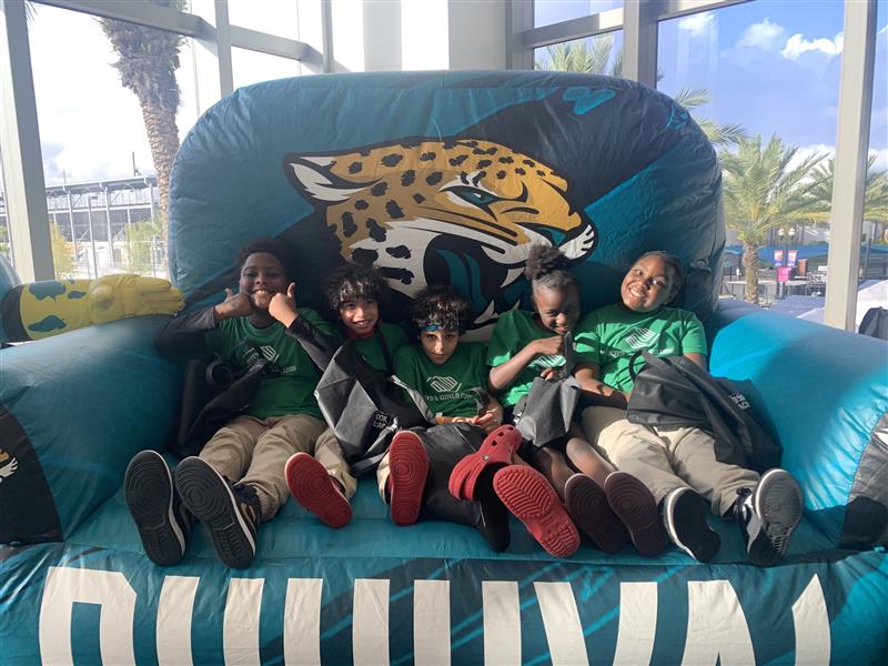 Five children in green shirts and khaki pants sitting on a large, inflatable blue chair with a Jacksonville Jaguars logo.
