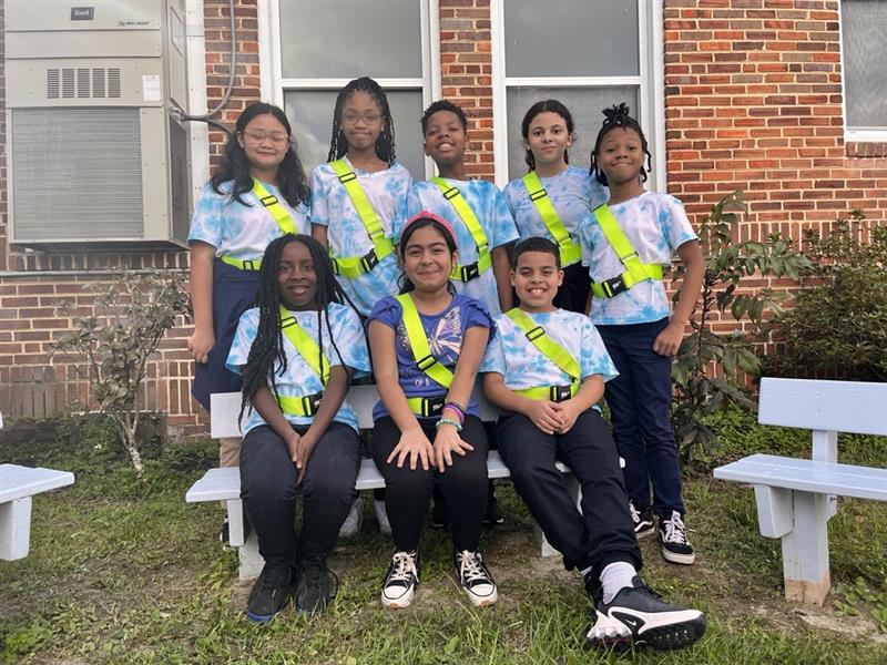 Group of young people wearing matching shirts with reflective yellow straps pose in front of a brick building.