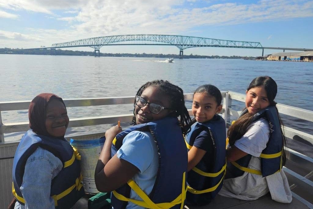 Four people in life vests sit on a boat deck, facing the camera. Behind them is a body of water and a bridge.