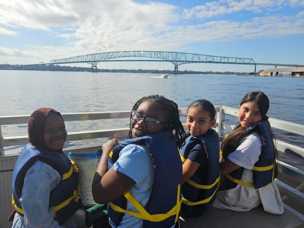 Four people in life jackets sit on a boat, with a bridge visible in the distance.