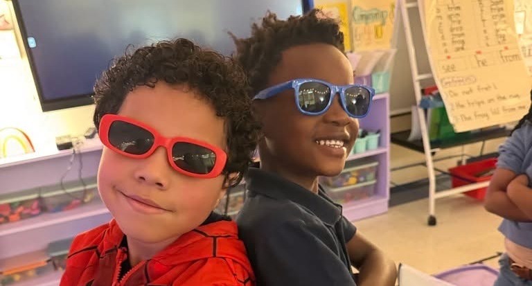 Two kids, one wearing red sunglasses and the other blue sunglasses, stand close together in a classroom.