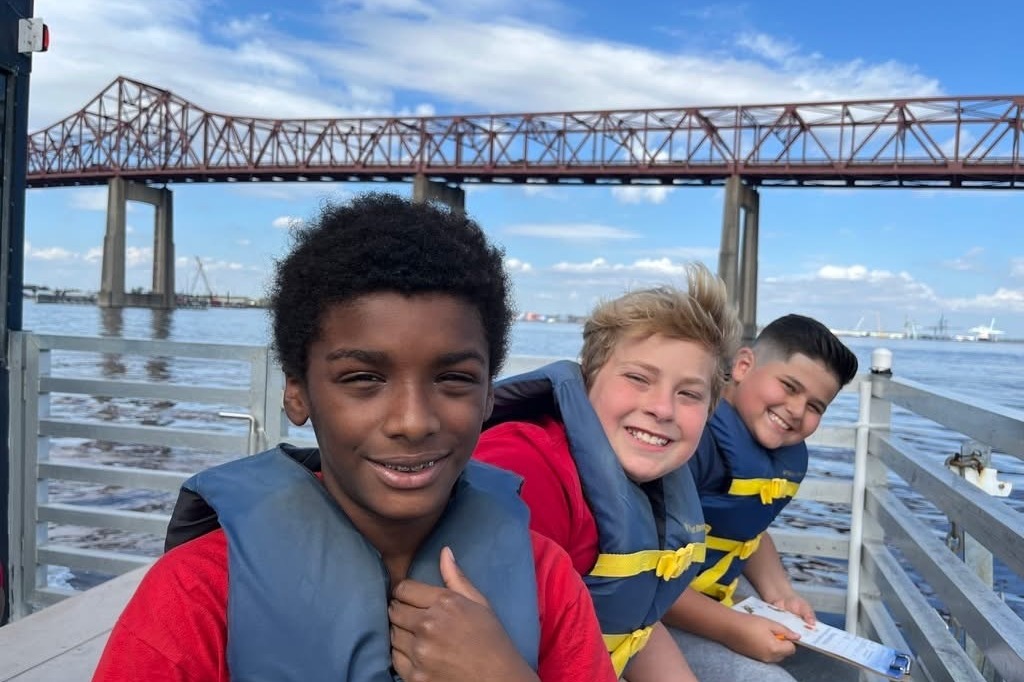 Three children in life jackets sit on a dock. Two wear red shirts, one has a blue shirt. Behind them, a bridge spans a body of water.
