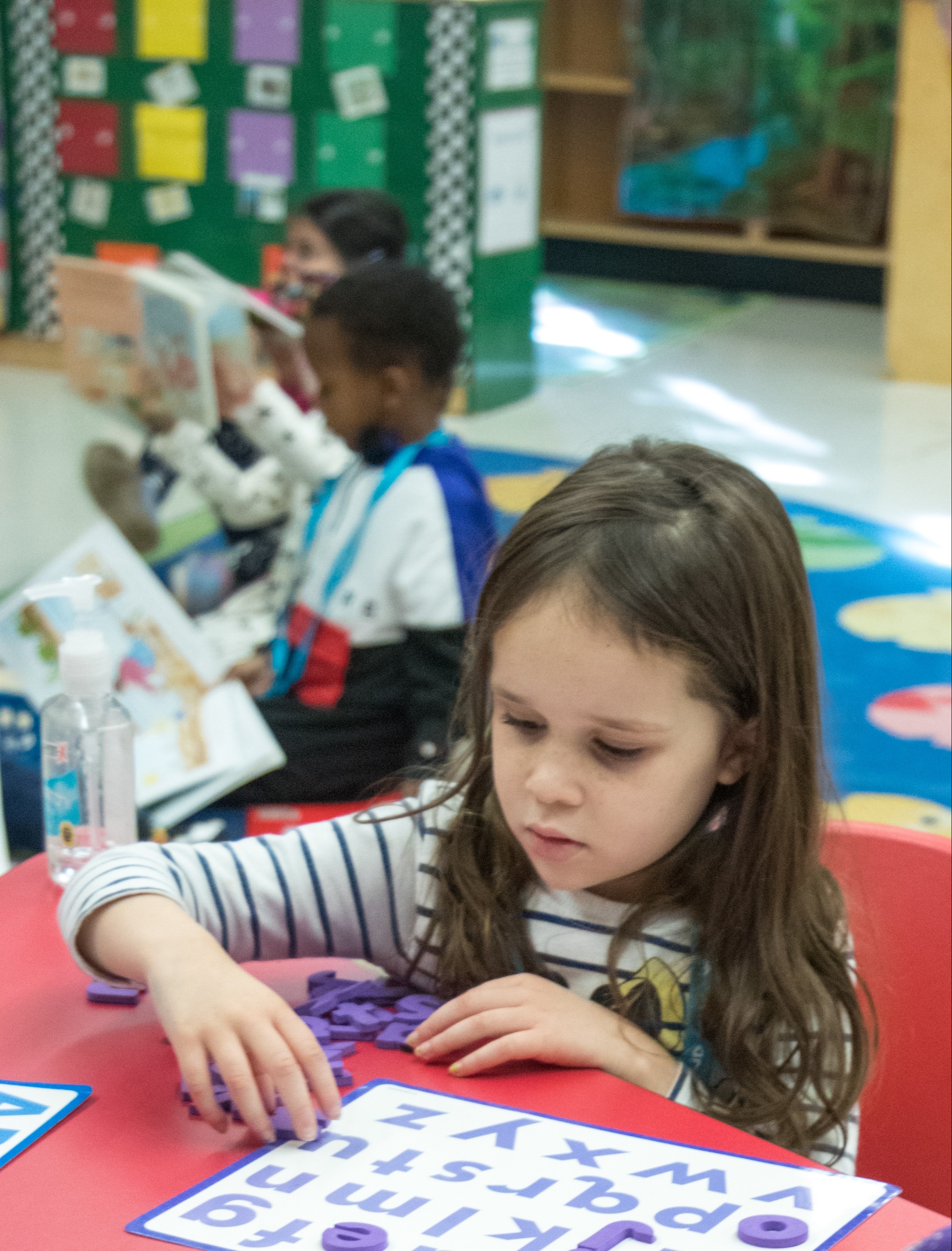 A girl with long hair sits at a table, concentrating on a word puzzle. Behind her, two children read books.