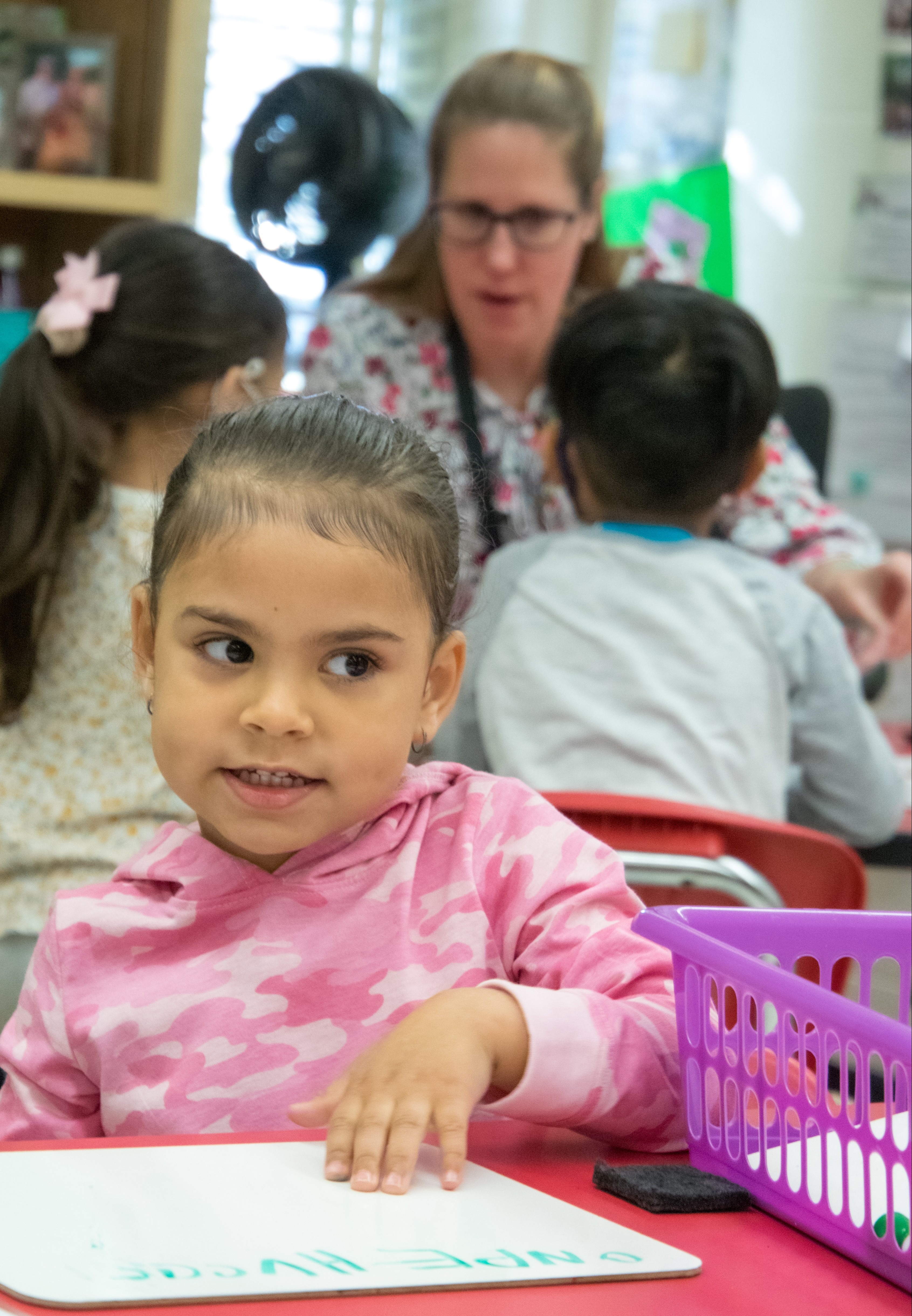 Girl in pink shirt seated at a table, writing on paper. Woman in glasses stands behind her. Background: shelves, chairs, and storage baskets.