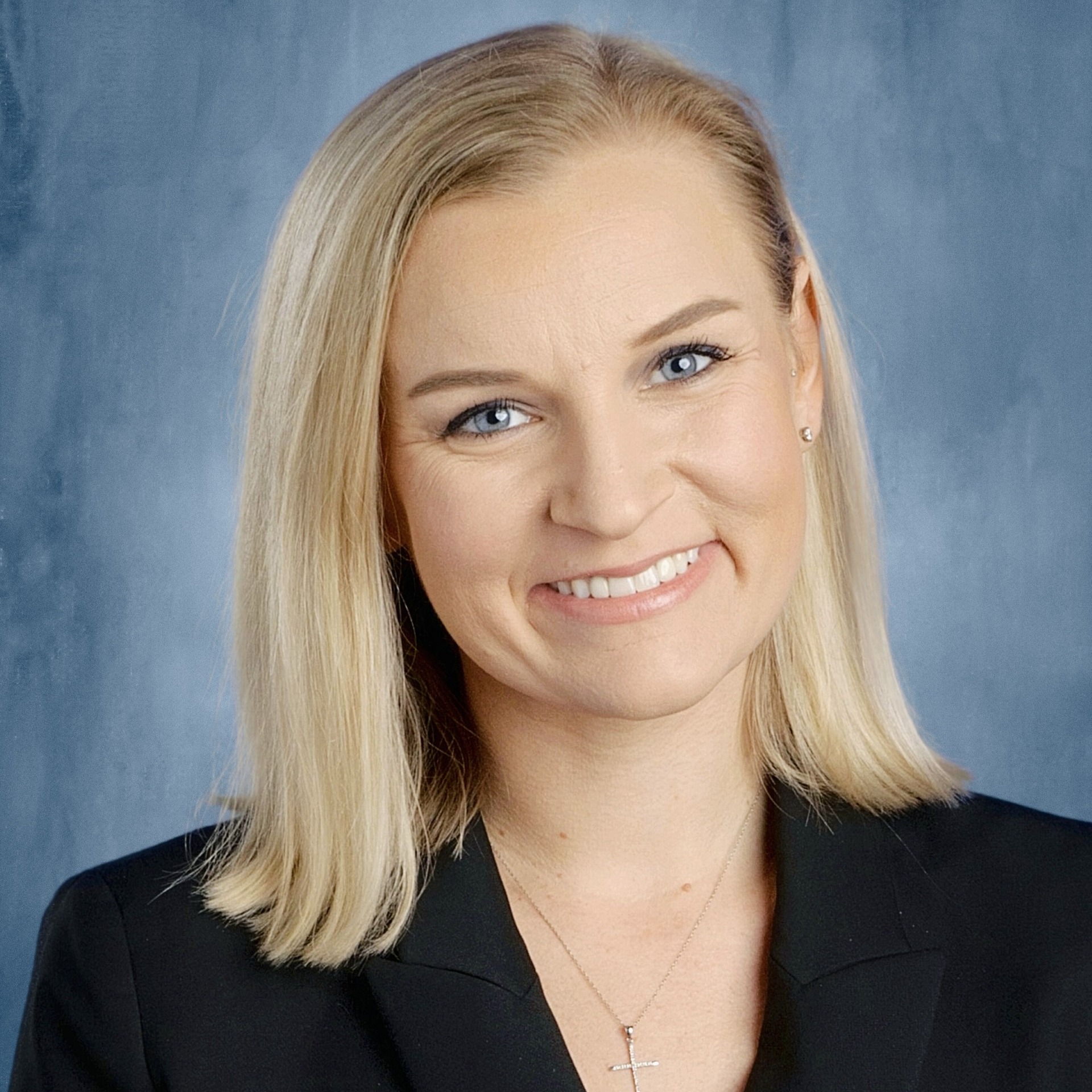A woman with blonde hair and blue eyes, wearing a black blazer, smiles at the camera.
