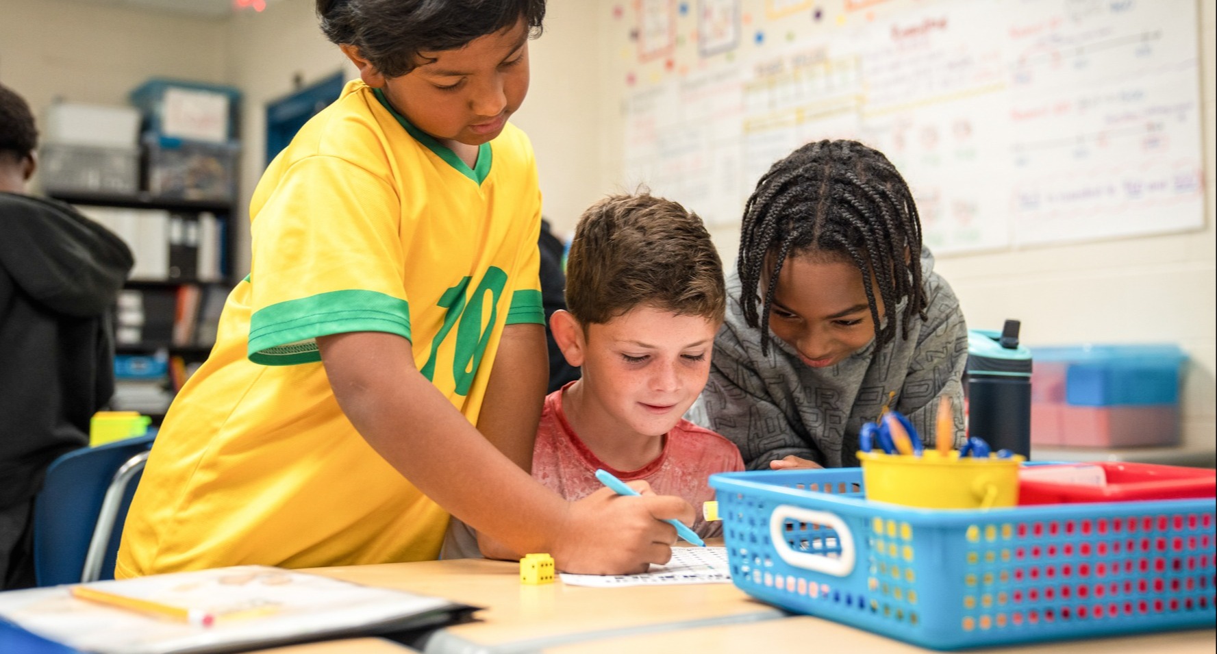 Three children at a desk. One leans over with a pencil. Another is seated, looking at a paper.
