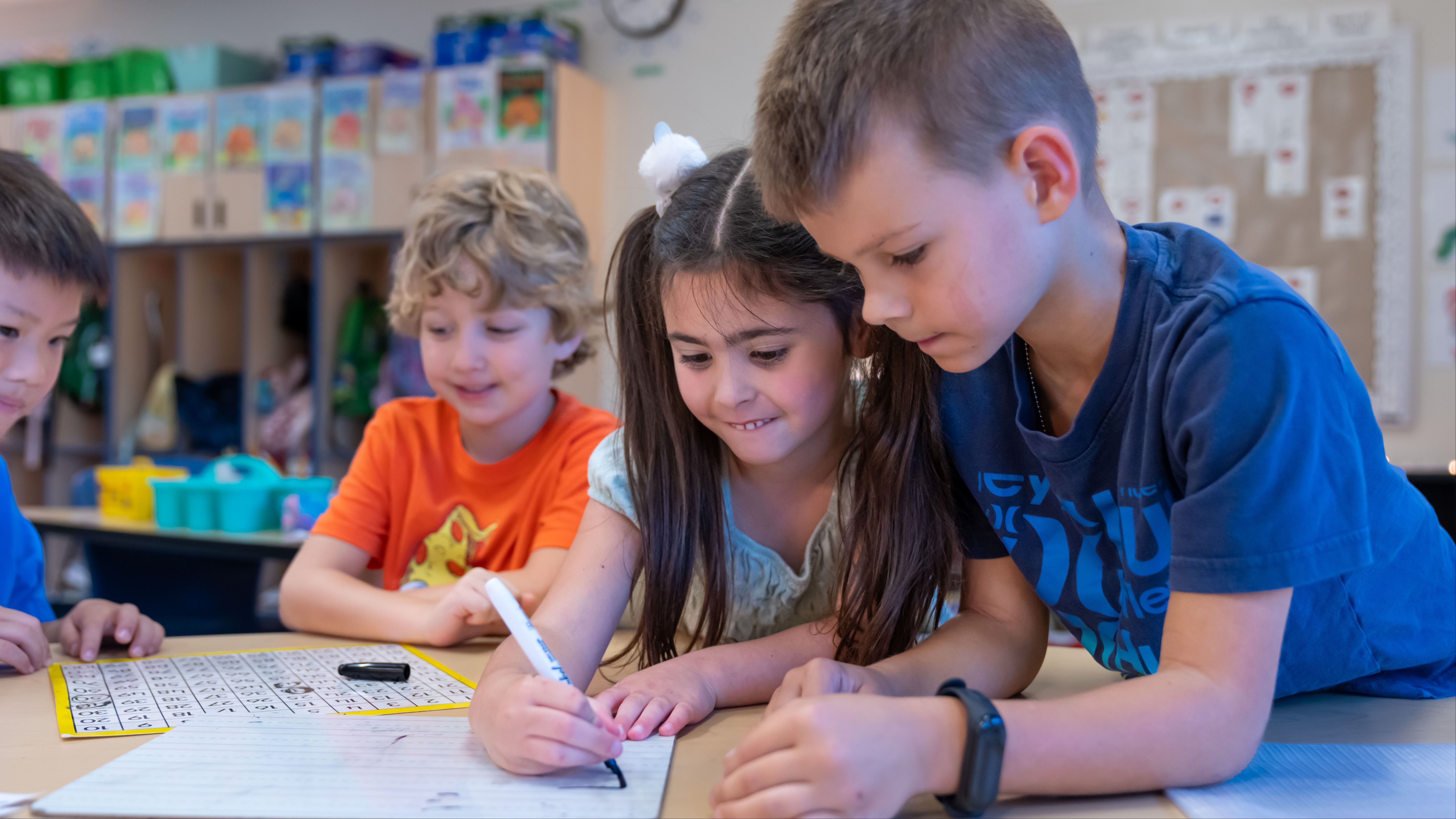 Four children seated around a table, using pens on paper. One child holds a marker.