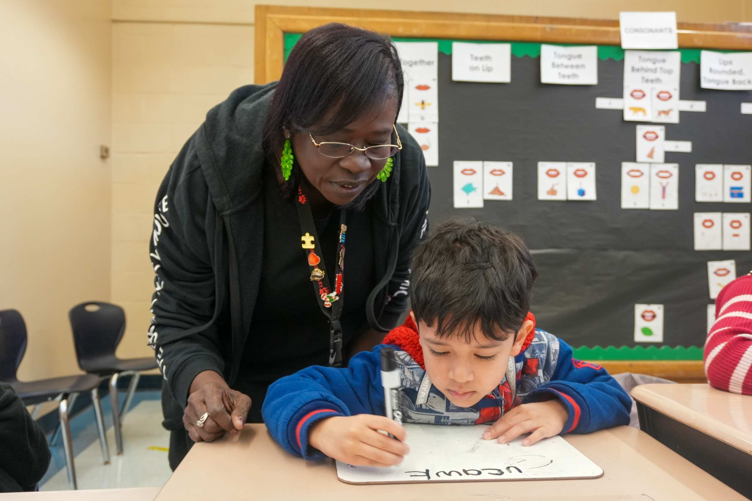 A student at a desk writing on a dry erase board, teacher looking over his shoulder helping him