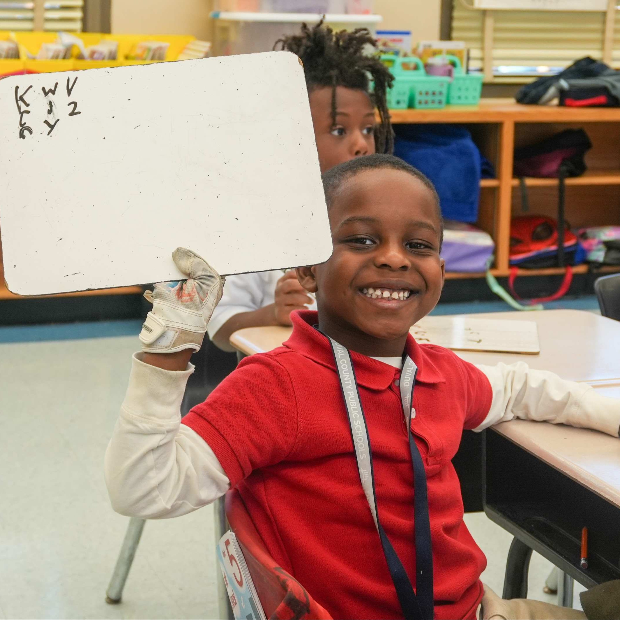 Child in a red shirt holding a whiteboard with letters while smiling in a classroom.