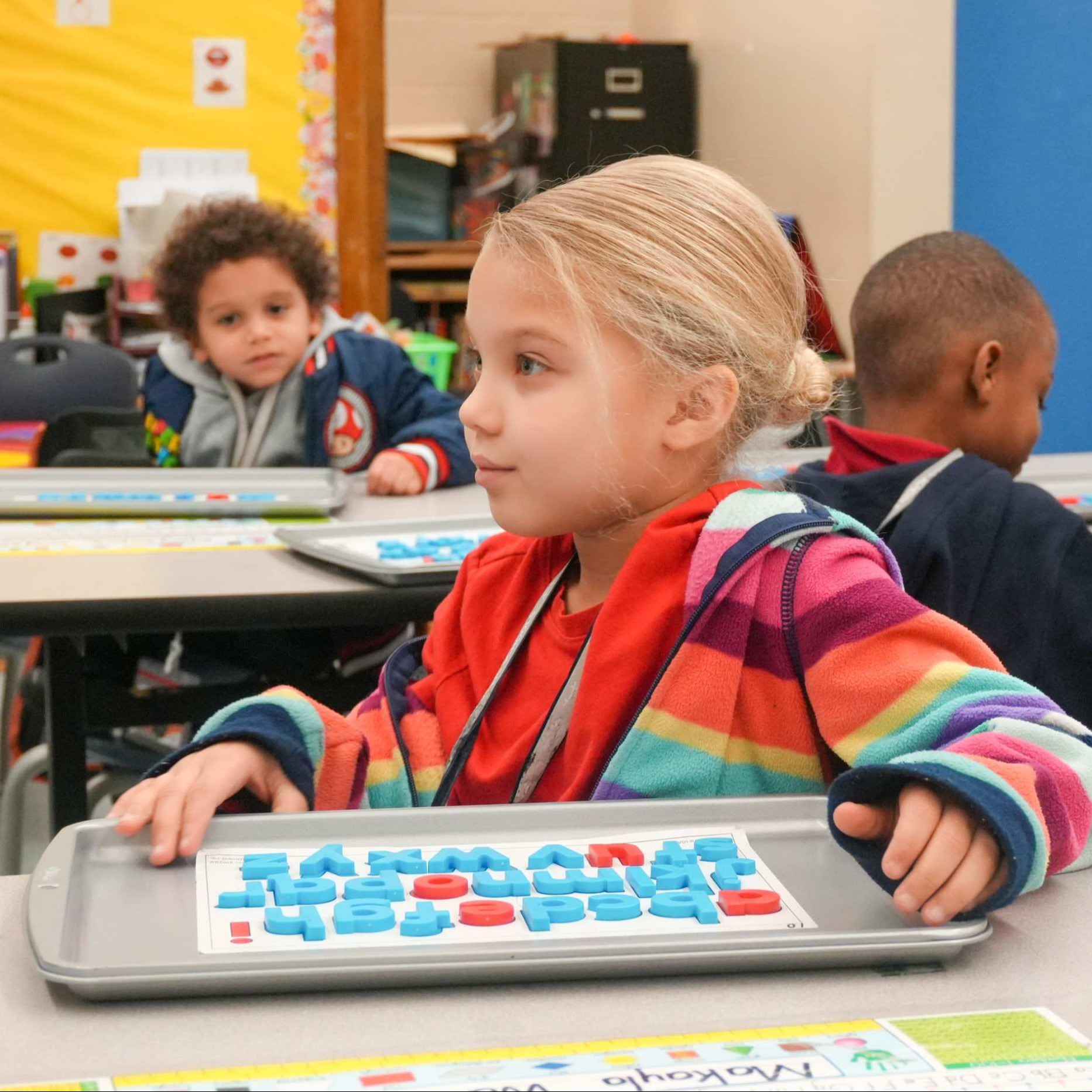 Child in a classroom uses a tactile alphabet board to learn. Behind, other students sit at desks.