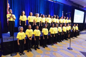 Children in yellow shirts stand in rows on stage, holding microphones and facing a projector screen.