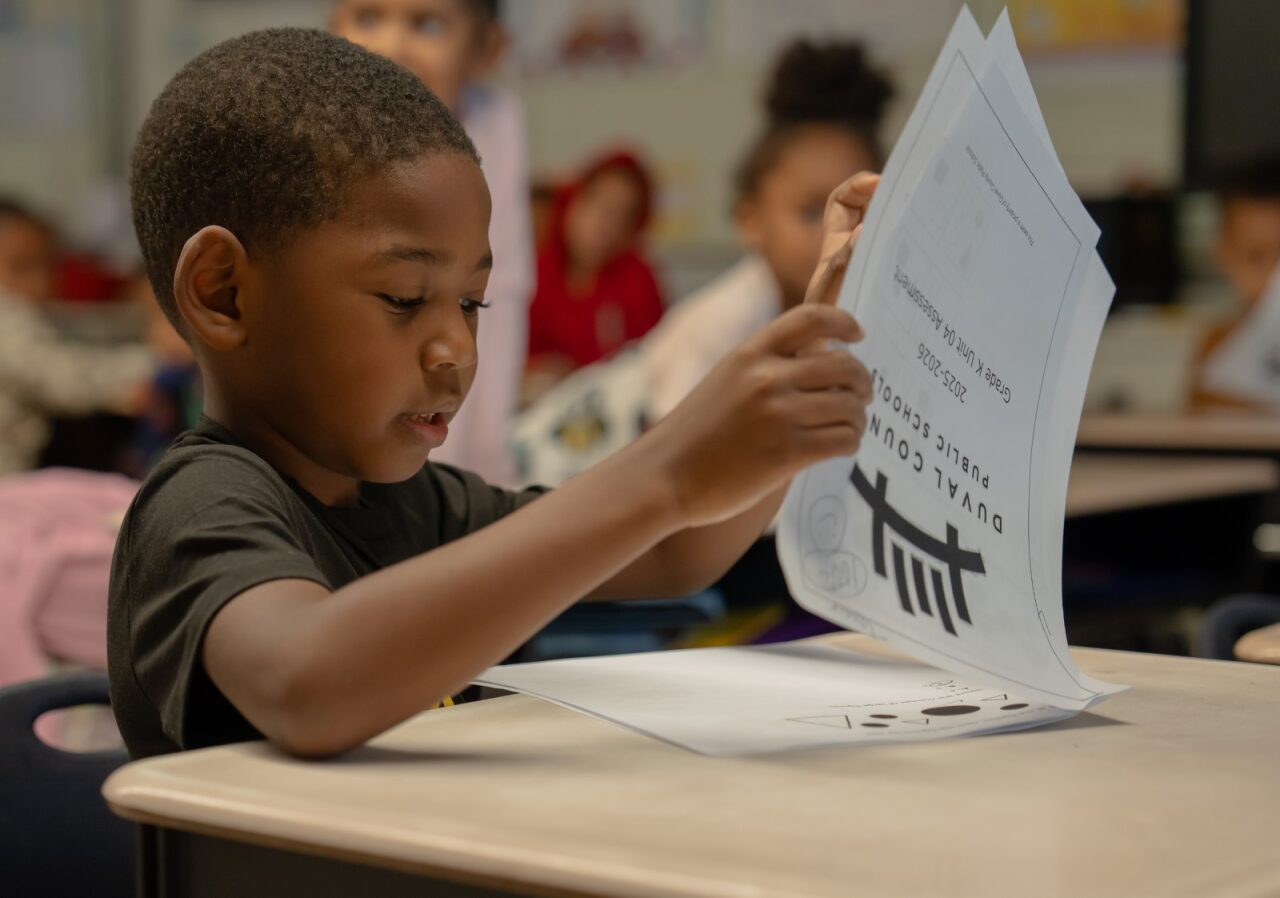 A boy reads a document in a classroom. Other students sit in the background, also engaged in reading.