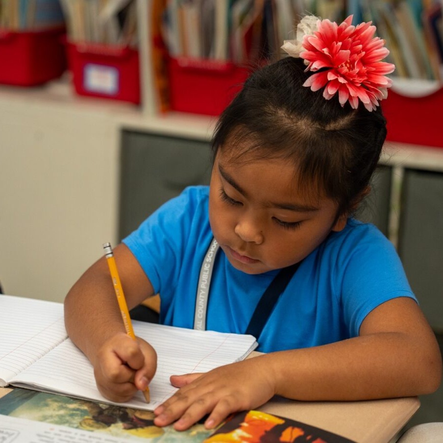 Child in a classroom, writing on a piece of paper with a pencil, and a colorful book on table.