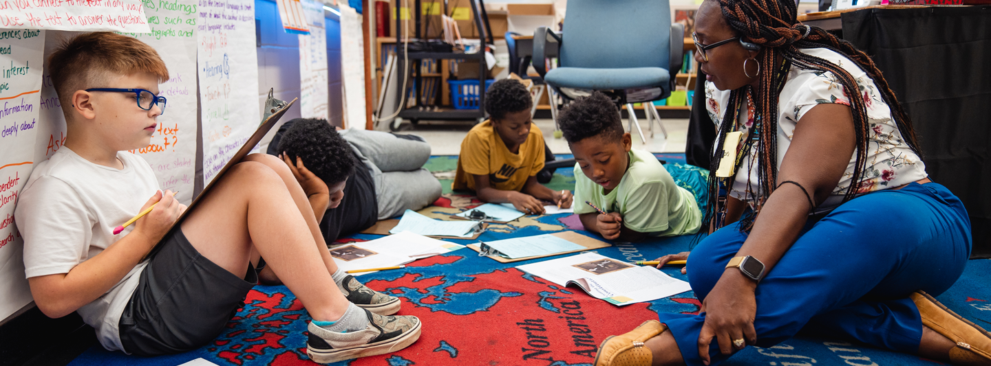 teacher sitting on floor with students writing in workbooks