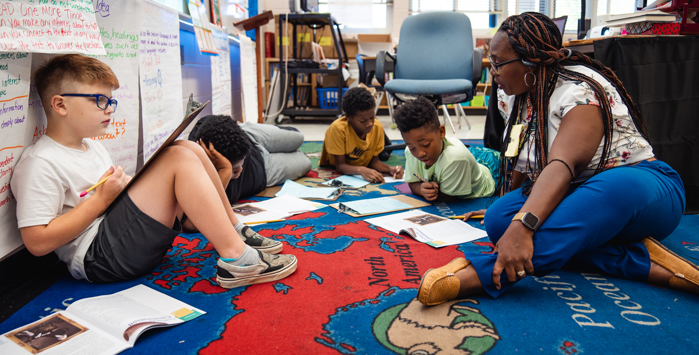 teacher sitting on floor with students writing in workbooks