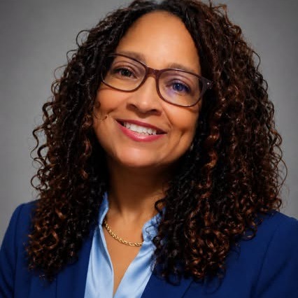 A woman with curly hair and glasses smiles in a blue suit and light blue shirt against a gray background.