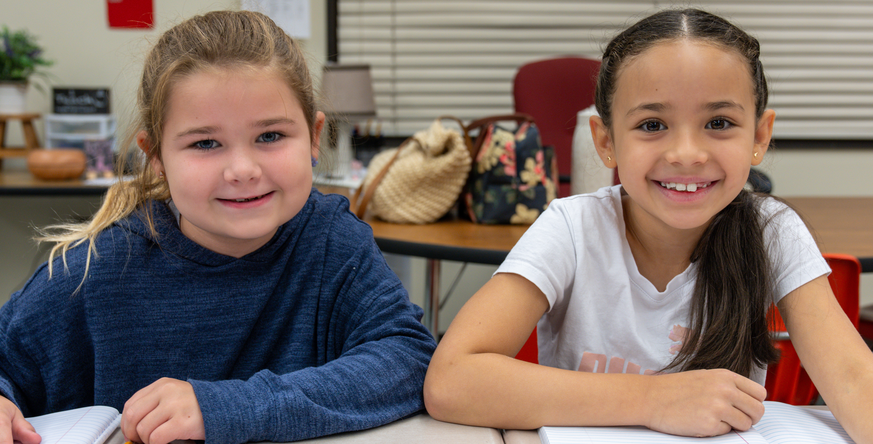 Two smiling young students, a girl with light brown hair and a girl with darker braided hair, sit together at a desk in a classroom.