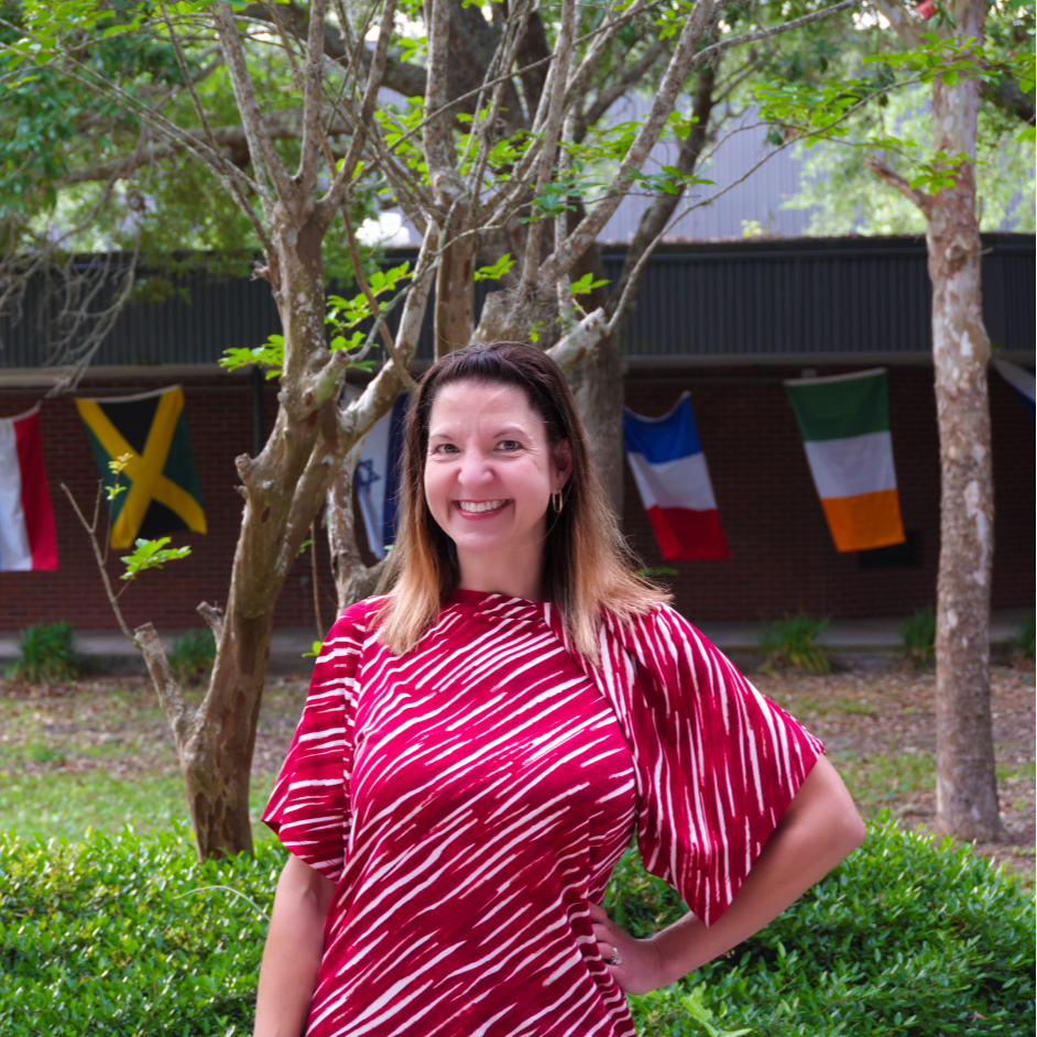A woman in a red and white striped dress smiles with her hands on her hips outside near trees and flags.