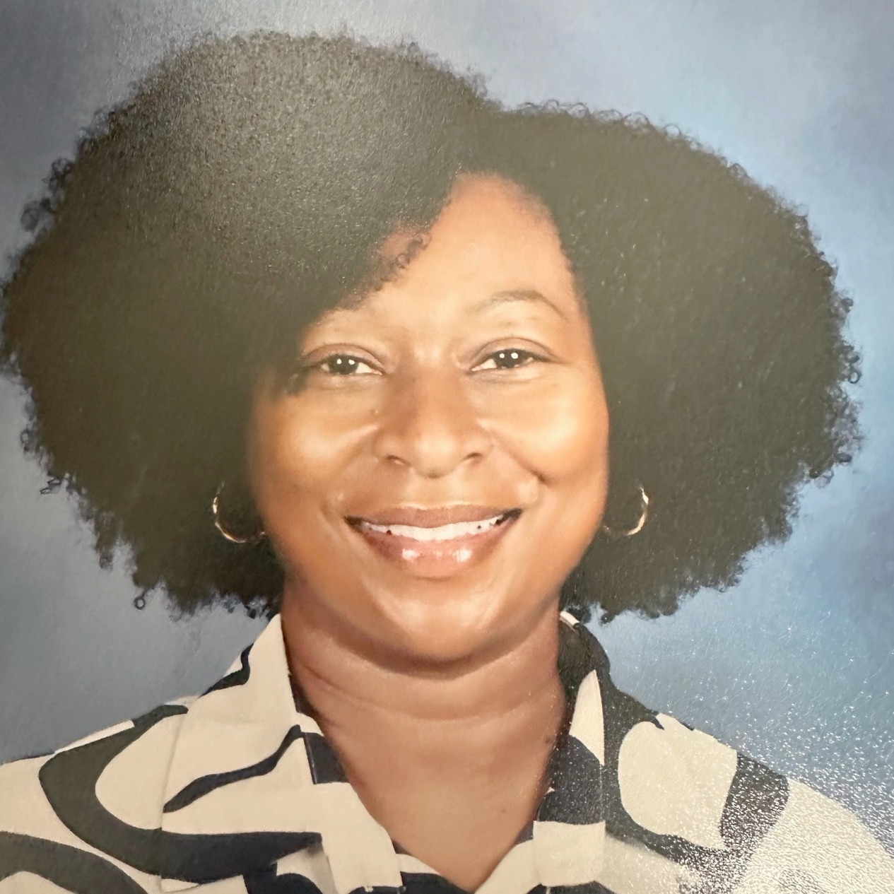 Person with short, curly hair wearing a white-and-black patterned collared shirt, smiling. Background is solid blue.