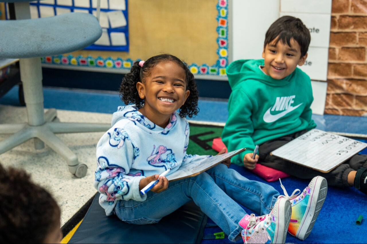 A girl smiling with a white board and market, a boy in the background looking at her and smiling