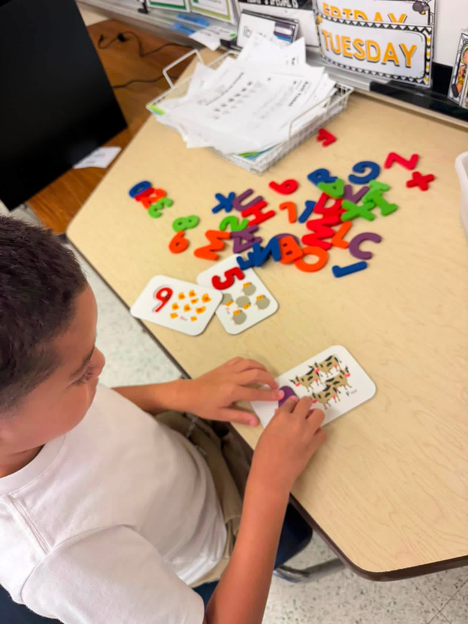 A young boy sits at a table, engaging with colorful alphabet tiles and number cards, seated in a classroom setting.